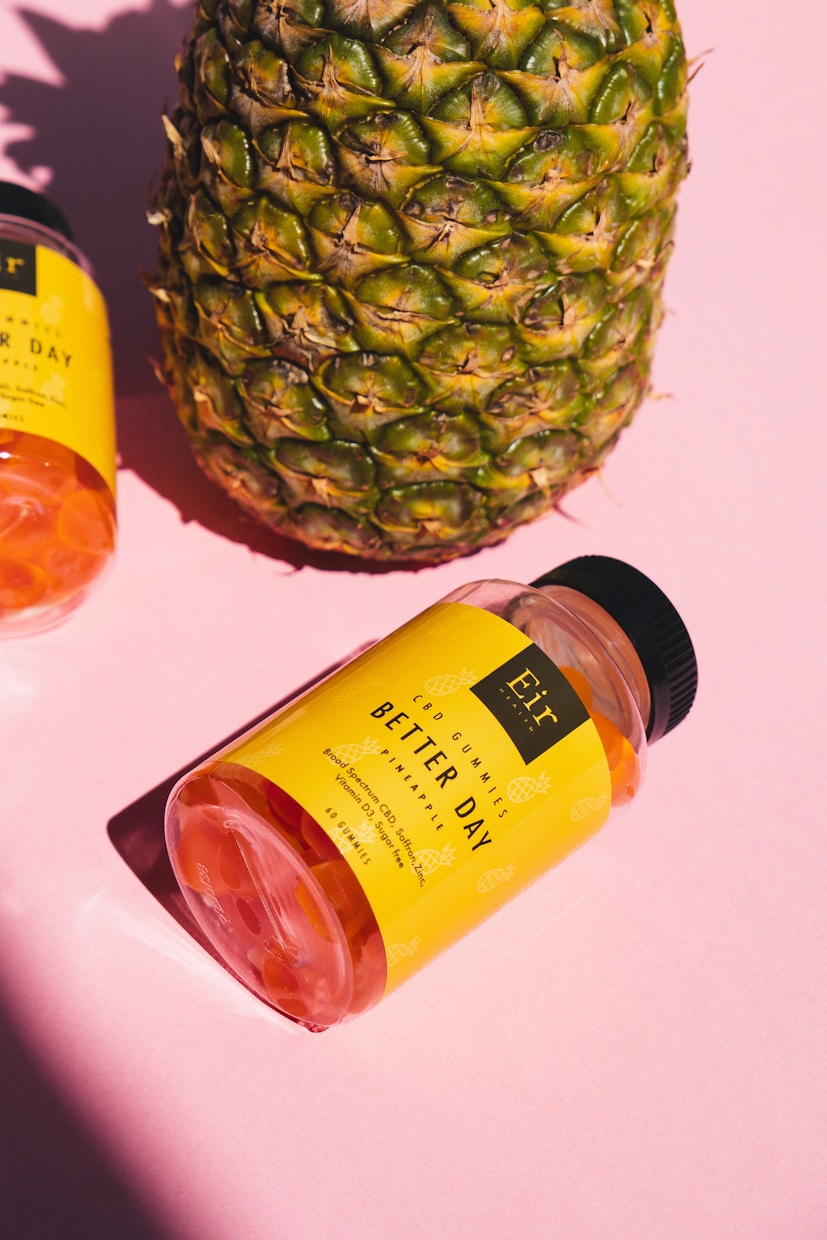 A large pineapple stands next to a bottle of gummy supplements labeled 'Better Day' on a pink background. The bottle has a bright yellow label and is tilted on its side.