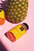 A large pineapple stands next to a bottle of gummy supplements labeled 'Better Day' on a pink background. The bottle has a bright yellow label and is tilted on its side.