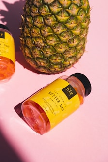 A large pineapple stands next to a bottle of gummy supplements labeled 'Better Day' on a pink background. The bottle has a bright yellow label and is tilted on its side.