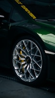 Close-up of a luxury sports car tire and rim with intricate design. The car is painted a dark green with a reflective finish. Yellow brake calipers are visible through the rim, and there are yellow letters on the windshield.