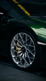Close-up of a luxury sports car tire and rim with intricate design. The car is painted a dark green with a reflective finish. Yellow brake calipers are visible through the rim, and there are yellow letters on the windshield.