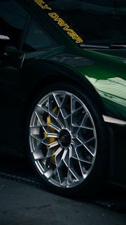 Close-up of a luxury sports car tire and rim with intricate design. The car is painted a dark green with a reflective finish. Yellow brake calipers are visible through the rim, and there are yellow letters on the windshield.