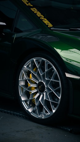 Close-up of a luxury sports car tire and rim with intricate design. The car is painted a dark green with a reflective finish. Yellow brake calipers are visible through the rim, and there are yellow letters on the windshield.