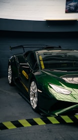 A sleek, high-performance sports car with a glossy dark green finish and a prominent rear spoiler is parked in an indoor garage. The car features custom decals, including yellow text on the windows and sides. The environment is slightly dim, highlighting the car's shiny surface and reflective qualities.