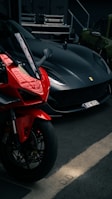A sleek black sports car is parked next to a vibrant red motorcycle in a dimly lit garage. The vehicles are positioned side by side, showcasing their curves and design details. In the background, there are some electrical equipment and a green car.