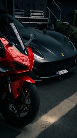 A sleek black sports car is parked next to a vibrant red motorcycle in a dimly lit garage. The vehicles are positioned side by side, showcasing their curves and design details. In the background, there are some electrical equipment and a green car.