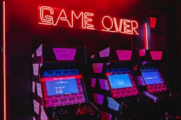 Neon red lighting illuminates a row of retro arcade machines, each displaying screens with pixelated graphics. The words 'GAME OVER' are prominently displayed in glowing red neon above the machines, creating a nostalgic and slightly eerie atmosphere. The machines are branded with 'House of Vans' stickers.