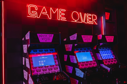 Neon red lighting illuminates a row of retro arcade machines, each displaying screens with pixelated graphics. The words 'GAME OVER' are prominently displayed in glowing red neon above the machines, creating a nostalgic and slightly eerie atmosphere. The machines are branded with 'House of Vans' stickers.