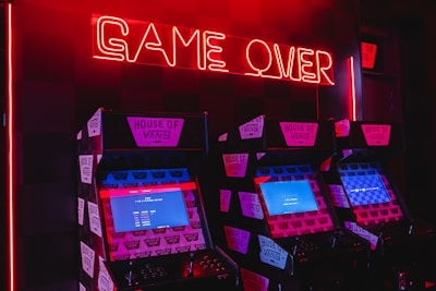 Neon red lighting illuminates a row of retro arcade machines, each displaying screens with pixelated graphics. The words 'GAME OVER' are prominently displayed in glowing red neon above the machines, creating a nostalgic and slightly eerie atmosphere. The machines are branded with 'House of Vans' stickers.