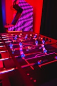 A foosball table with colorful players is illuminated by vibrant red and purple lighting, creating a lively and playful atmosphere. In the background, there is an arcade machine with a geometric pattern.