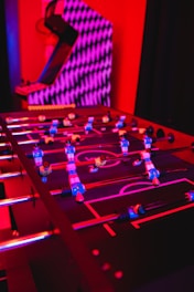 A foosball table with colorful players is illuminated by vibrant red and purple lighting, creating a lively and playful atmosphere. In the background, there is an arcade machine with a geometric pattern.