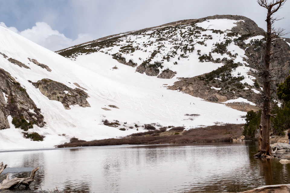 The image depicts a serene winter landscape with a mountain covered in snow and a lake nestled at its base, reflecting the surrounding scenery. The sky above is overcast, casting a soft light on the scene below. On the right side of the image, there are trees standing tall against the snowy backdrop, adding depth to the composition.

In the foreground, there's a small body of water that mirrors the mountain and sky, creating a beautiful reflection. The lake appears calm with no visible ripples or waves. The overall scene is peaceful and tranquil, showcasing the beauty of nature during winter.