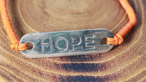 A metallic plate engraved with the word 'HOPE' is attached to an orange cord, forming a bracelet. The bracelet is placed on a surface with a wood-like pattern, highlighting the texture and color contrast between the metal, cord, and background.