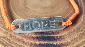 A metallic plate engraved with the word 'HOPE' is attached to an orange cord, forming a bracelet. The bracelet is placed on a surface with a wood-like pattern, highlighting the texture and color contrast between the metal, cord, and background.