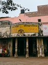 An old, weathered building with faded yellow signage in Hindi, indicating it's part of the Uttar Pradesh Transport Corporation bus station in a place called Sambhal. The structure shows signs of aging, with peeling paint and visible grime. Two women, dressed in traditional attire, sit on a bench outside the building. A pink wall and a black water tank are visible in the background, with an overhanging tree branch partially in view.
