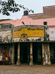 An old, weathered building with faded yellow signage in Hindi, indicating it's part of the Uttar Pradesh Transport Corporation bus station in a place called Sambhal. The structure shows signs of aging, with peeling paint and visible grime. Two women, dressed in traditional attire, sit on a bench outside the building. A pink wall and a black water tank are visible in the background, with an overhanging tree branch partially in view.