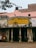 An old, weathered building with faded yellow signage in Hindi, indicating it's part of the Uttar Pradesh Transport Corporation bus station in a place called Sambhal. The structure shows signs of aging, with peeling paint and visible grime. Two women, dressed in traditional attire, sit on a bench outside the building. A pink wall and a black water tank are visible in the background, with an overhanging tree branch partially in view.