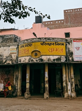 An old, weathered building with faded yellow signage in Hindi, indicating it's part of the Uttar Pradesh Transport Corporation bus station in a place called Sambhal. The structure shows signs of aging, with peeling paint and visible grime. Two women, dressed in traditional attire, sit on a bench outside the building. A pink wall and a black water tank are visible in the background, with an overhanging tree branch partially in view.