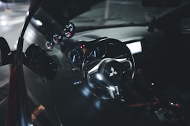 The interior of a car featuring a close-up view of the steering wheel with a distinctive logo in the center. The dashboard has illuminated gauges, providing a modern and sleek appearance. The reflection of the interior is visible on the side window, adding depth to the image.