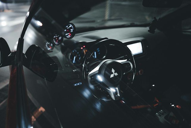 The interior of a car featuring a close-up view of the steering wheel with a distinctive logo in the center. The dashboard has illuminated gauges, providing a modern and sleek appearance. The reflection of the interior is visible on the side window, adding depth to the image.