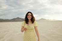 A smiling woman wearing a bright yellow dress standing in a sunlit city street.
