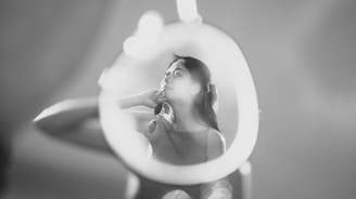 A woman gazes pensively at her reflection in a mirror. The scene is captured in black and white, with a soft, dreamy focus. The lighting creates a serene and contemplative atmosphere.