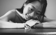 A woman with a gentle smile gazes intently at a large diamond placed on a table. The scene is captured in black and white, highlighting the contrast between the dark backdrop and the sparkling facets of the diamond.