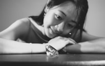 A woman with a gentle smile gazes intently at a large diamond placed on a table. The scene is captured in black and white, highlighting the contrast between the dark backdrop and the sparkling facets of the diamond.