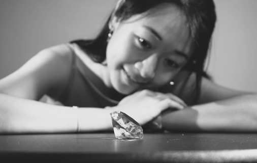 A woman with a gentle smile gazes intently at a large diamond placed on a table. The scene is captured in black and white, highlighting the contrast between the dark backdrop and the sparkling facets of the diamond.