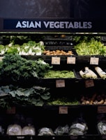 A grocery store display featuring a variety of Asian vegetables. The neatly arranged produce includes leafy greens such as bok choy and napa cabbage, as well as other items like ginger and daikon radishes. Price tags are visible, indicating different prices for the vegetables.