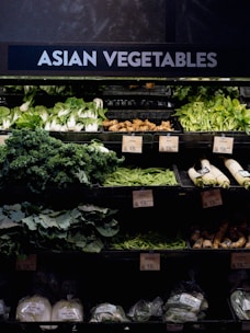 A grocery store display featuring a variety of Asian vegetables. The neatly arranged produce includes leafy greens such as bok choy and napa cabbage, as well as other items like ginger and daikon radishes. Price tags are visible, indicating different prices for the vegetables.