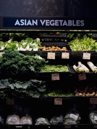 A grocery store display featuring a variety of Asian vegetables. The neatly arranged produce includes leafy greens such as bok choy and napa cabbage, as well as other items like ginger and daikon radishes. Price tags are visible, indicating different prices for the vegetables.