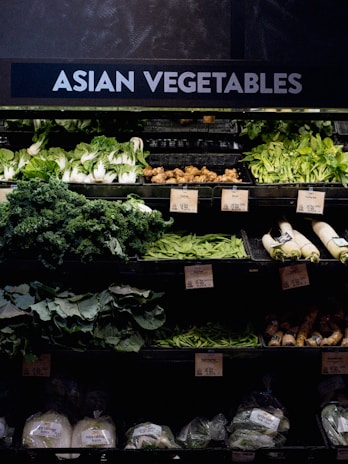 A grocery store display featuring a variety of Asian vegetables. The neatly arranged produce includes leafy greens such as bok choy and napa cabbage, as well as other items like ginger and daikon radishes. Price tags are visible, indicating different prices for the vegetables.