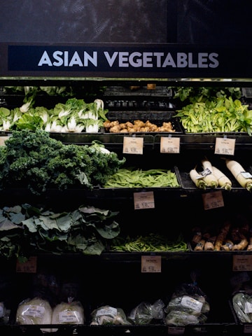 A grocery store display featuring a variety of Asian vegetables. The neatly arranged produce includes leafy greens such as bok choy and napa cabbage, as well as other items like ginger and daikon radishes. Price tags are visible, indicating different prices for the vegetables.