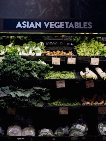 A grocery store display featuring a variety of Asian vegetables. The neatly arranged produce includes leafy greens such as bok choy and napa cabbage, as well as other items like ginger and daikon radishes. Price tags are visible, indicating different prices for the vegetables.