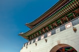 The image features a traditional Korean architectural structure with ornate detailing and vibrant colors. The roof showcases intricate green and red patterns with white and orange accents. Stone elements with carved designs are visible, and the sky is a clear, deep blue.