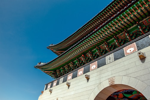 The image features a traditional Korean architectural structure with ornate detailing and vibrant colors. The roof showcases intricate green and red patterns with white and orange accents. Stone elements with carved designs are visible, and the sky is a clear, deep blue.