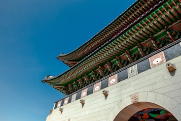 The image features a traditional Korean architectural structure with ornate detailing and vibrant colors. The roof showcases intricate green and red patterns with white and orange accents. Stone elements with carved designs are visible, and the sky is a clear, deep blue.