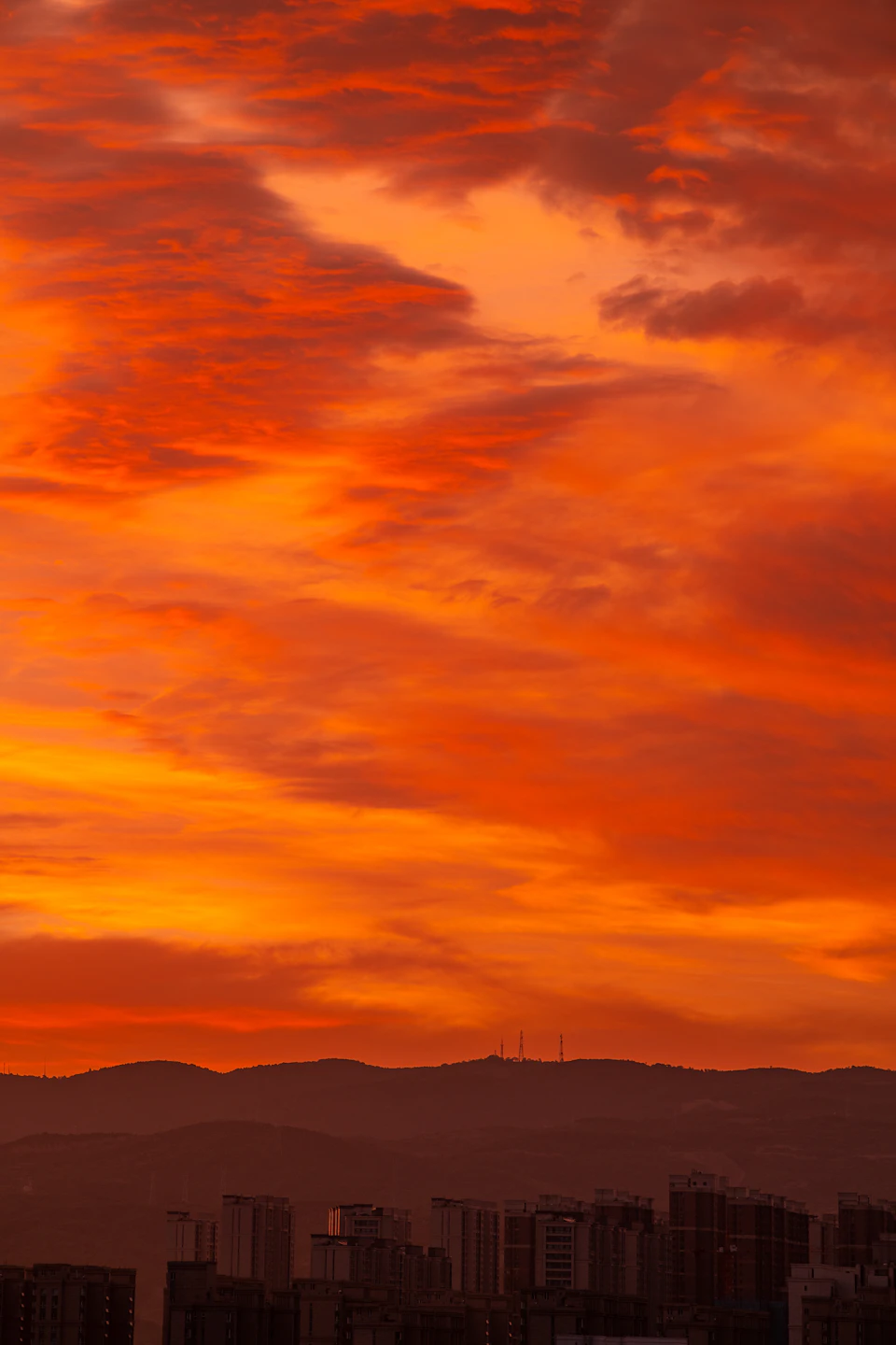The image depicts a breathtaking sunset over a cityscape, with an orange sky serving as a stunning backdrop to the urban landscape below. The sun is setting behind the buildings, casting long shadows and bathing the scene in warm hues. The perspective from which the photo was taken allows for a comprehensive view of the city's skyline, with numerous tall buildings stretching towards the horizon. The colors in the image are vibrant, with the orange sky contrasting beautifully against the silhouettes of the buildings.