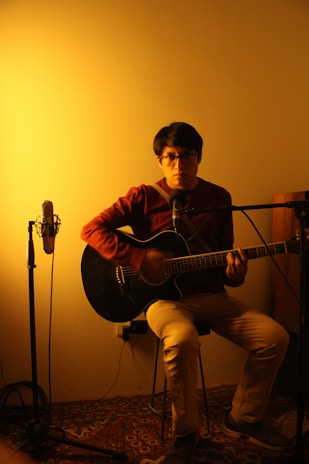 A musician playing guitar in a cozy recording booth surrounded by microphones.