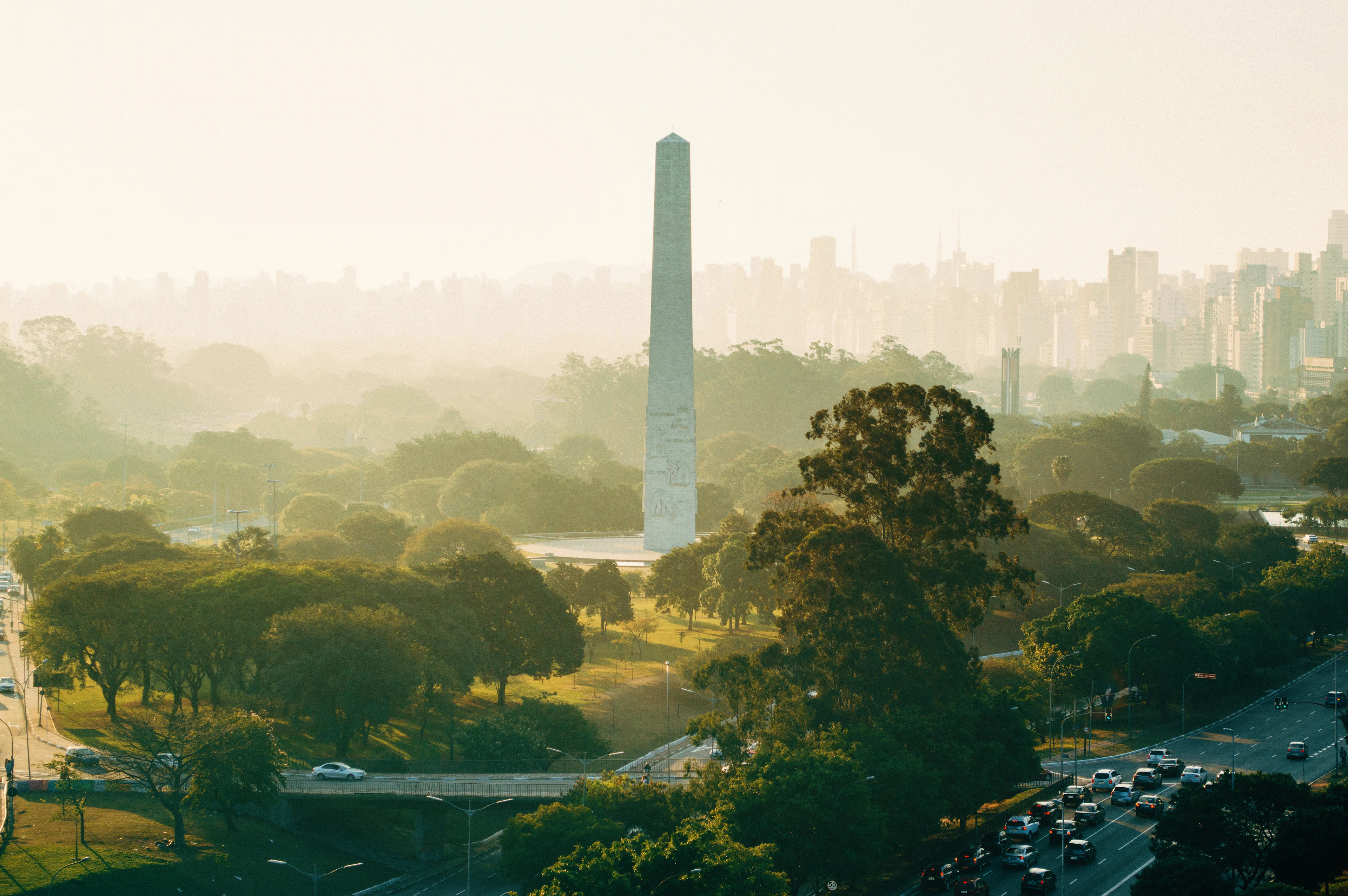 The white Obelisco de São Paulo rising out of the tree canopy of Ibirapuera Park in low golden haze, with the city's high-rise skyline dissolving into mist behind it and traffic threading the park road below