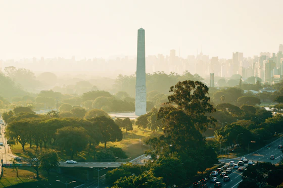 Obelisco do Ibirapuera em área gramada do Parque do Ibirapuera, com a linha de prédios de São Paulo ao fundo na neblina