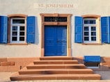 A building facade features a central blue door with decorative brickwork framing and two windows on either side. Each window has functional blue shutters, and there are steps leading up to the door. Above the door, a sign reads 'ST. JOSEFSHEIM'.