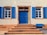 A building facade features a central blue door with decorative brickwork framing and two windows on either side. Each window has functional blue shutters, and there are steps leading up to the door. Above the door, a sign reads 'ST. JOSEFSHEIM'.
