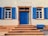 A building facade features a central blue door with decorative brickwork framing and two windows on either side. Each window has functional blue shutters, and there are steps leading up to the door. Above the door, a sign reads 'ST. JOSEFSHEIM'.