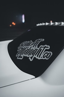 A dark car window featuring a decal with stylized text 'Ride At Midnight.co' in white. The decal stands out against the tinted rear windshield of the vehicle. The background is dimly lit with hints of distant blurred lights adding a subdued ambiance.