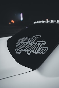 A dark car window featuring a decal with stylized text 'Ride At Midnight.co' in white. The decal stands out against the tinted rear windshield of the vehicle. The background is dimly lit with hints of distant blurred lights adding a subdued ambiance.