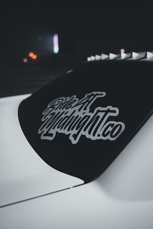 A dark car window featuring a decal with stylized text 'Ride At Midnight.co' in white. The decal stands out against the tinted rear windshield of the vehicle. The background is dimly lit with hints of distant blurred lights adding a subdued ambiance.