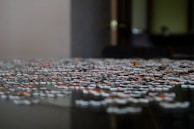 A close-up of a jigsaw puzzle on a table, with pieces scattered and some partially connected. The background is blurred, giving a focused perspective on the puzzle pieces, which display various colors and patterns.