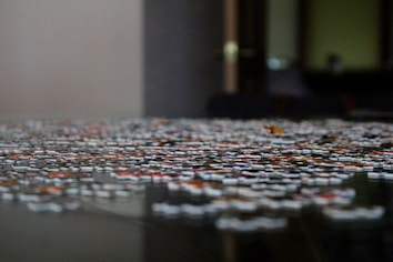 A close-up of a jigsaw puzzle on a table, with pieces scattered and some partially connected. The background is blurred, giving a focused perspective on the puzzle pieces, which display various colors and patterns.